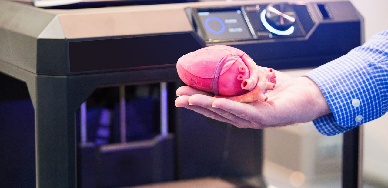 A hand holds a 3D printed heart in front of a 3D printer.